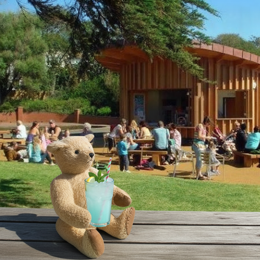 A plush teddy bear sits on a wooden deck holding a light blue drink with a green straw, with an outdoor cafe, people, and trees in the background.