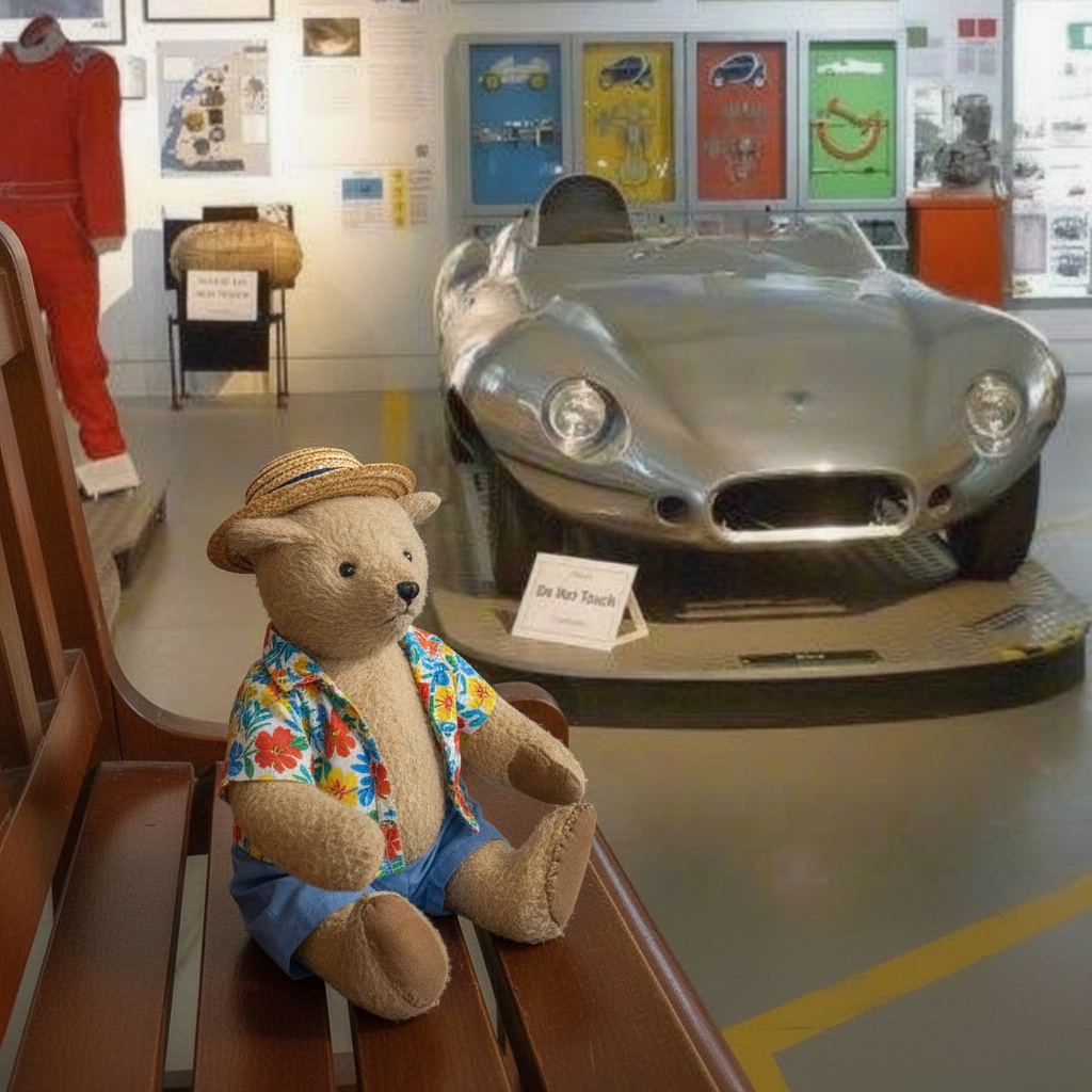 A teddy bear wearing a straw hat, colorful floral shirt, and blue shorts sits on a wooden bench in a museum display area, with a silver race car and exhibits in the background.